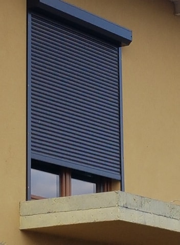 Zamknięta, grafitowa roleta zewnętrzna na oknie z drewnianą ramą, zamontowana na żółtej elewacji budynku z betonowym parapetem.