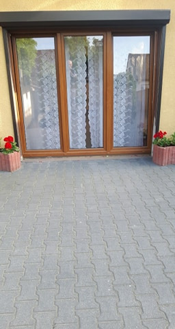 Trzyczęściowe okno z brązową ramą i roletą zewnętrzną, ozdobione koronkowymi firankami i doniczkami z czerwonymi kwiatami po bokach, widok z zewnątrz na brukowany podjazd.
