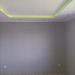 Bałabuch - Pomalowane na szaro wnętrze pokoju z podwieszanym sufitem z oświetleniem LED w kolorze żółtym, jasna podłoga z paneli.