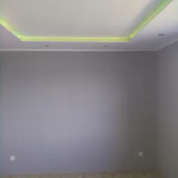 Pomalowane na szaro wnętrze pokoju z podwieszanym sufitem z oświetleniem LED w kolorze żółtym, jasna podłoga z paneli.