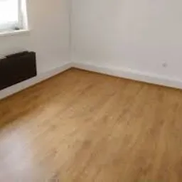 Pusty pokój po ułożeniu paneli podłogowych w odcieniach brązu; widoczna listwa przypodłogowa i grzejnik pod oknem.