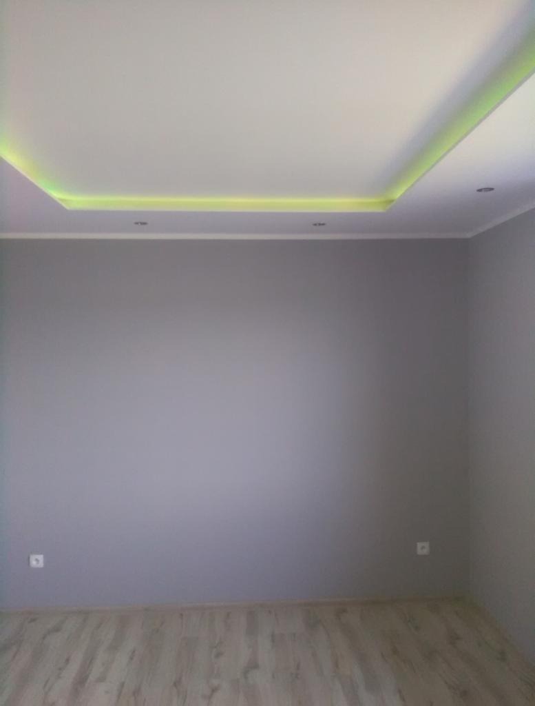 Pomalowane na szaro wnętrze pokoju z podwieszanym sufitem z oświetleniem LED w kolorze żółtym, jasna podłoga z paneli.