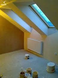 Poddasze po malowaniu, widoczne okna dachowe, grzejnik, puszki z farbą na podłodze, ściany w odcieniach żółci i beżu, surowe wykończenie podłogi.