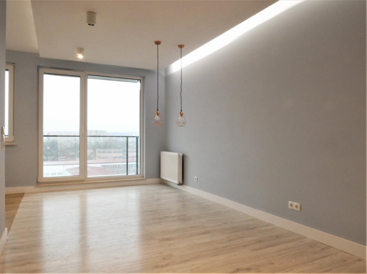 Pomalowane na szaro wnętrze pokoju z panelami podłogowymi, białymi listwami przypodłogowymi, grzejnikiem, dwoma lampami wiszącymi w miedzianych kloszach i dużym oknem balkonowym.