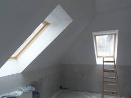 Poddasze po świeżym malowaniu na biało, widoczne dwa okna dachowe o różnej wielkości, drewniana drabina oparta o ścianę przy mniejszym oknie, ślady niedomalowań i zabrudzeń na ścianach.