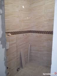 Wykończona ściana łazienki z beżowych płytek imitujących drewno, z dekoracyjnym pasem mozaiki w odcieniach brązu, widoczne narzędzia i resztki materiałów budowlanych.