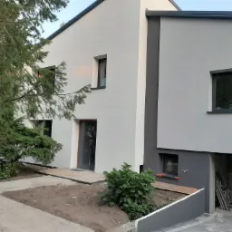 Nowoczesny, dwukondygnacyjny dom z jasnoszarą elewacją, ciemnoszarą stolarką okienną i drzwiami, widoczny fragment ogrodu z drzewem iglastym i utwardzonym podjazdem, stan surowy otwarty garażu.