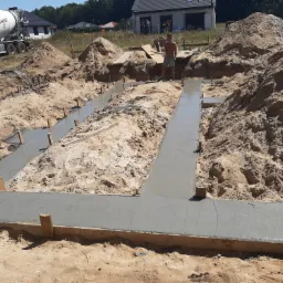 Wylane ławy fundamentowe na piaszczystym gruncie, z widocznym pracownikiem i betoniarką w tle.