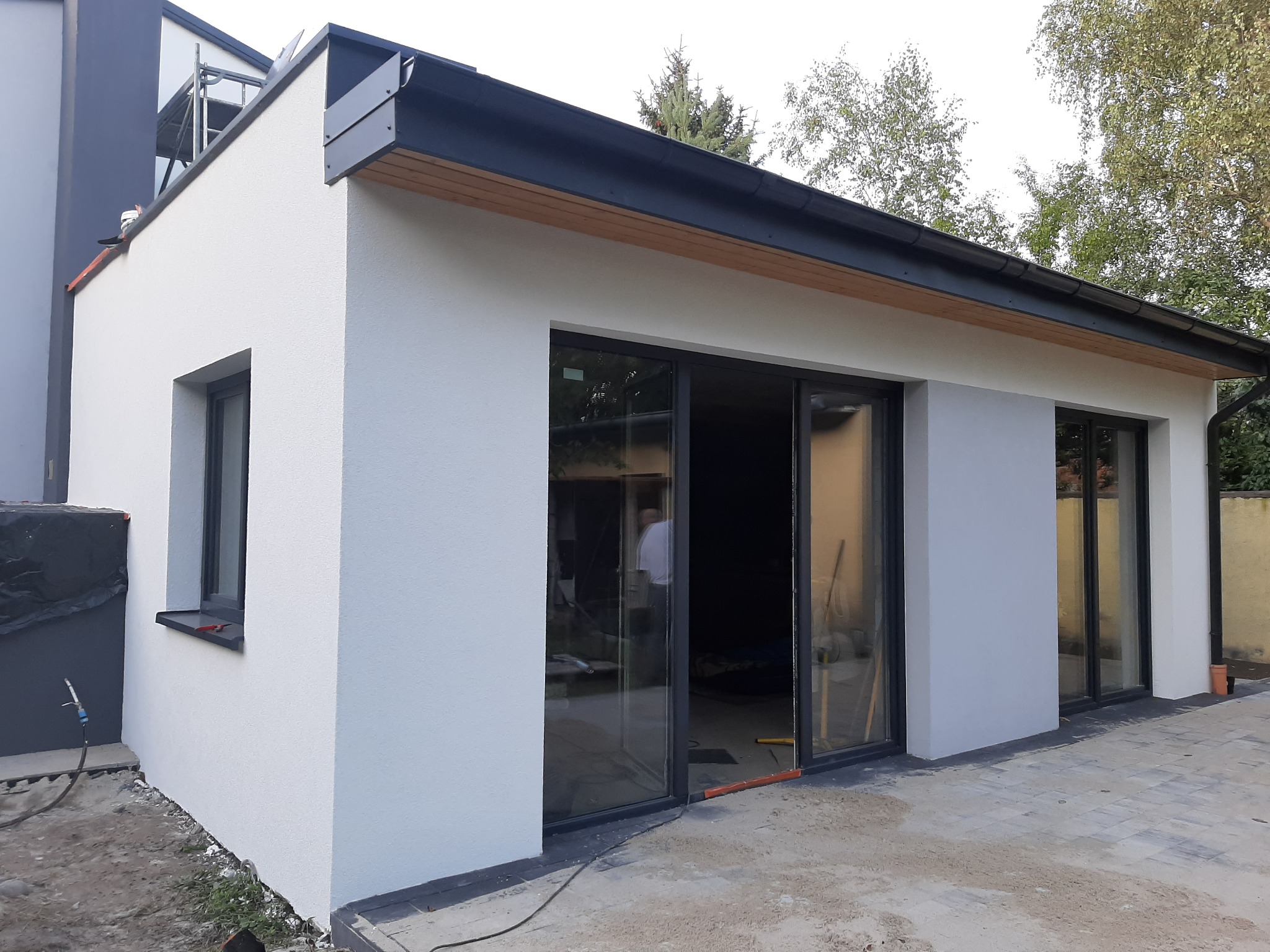 Nowoczesna dobudówka z dużymi przesuwnymi oknami i świeżo otynkowaną elewacją w kolorze jasnej szarości, widoczne elementy wykończeniowe dachu i kostka brukowa na zewnątrz.