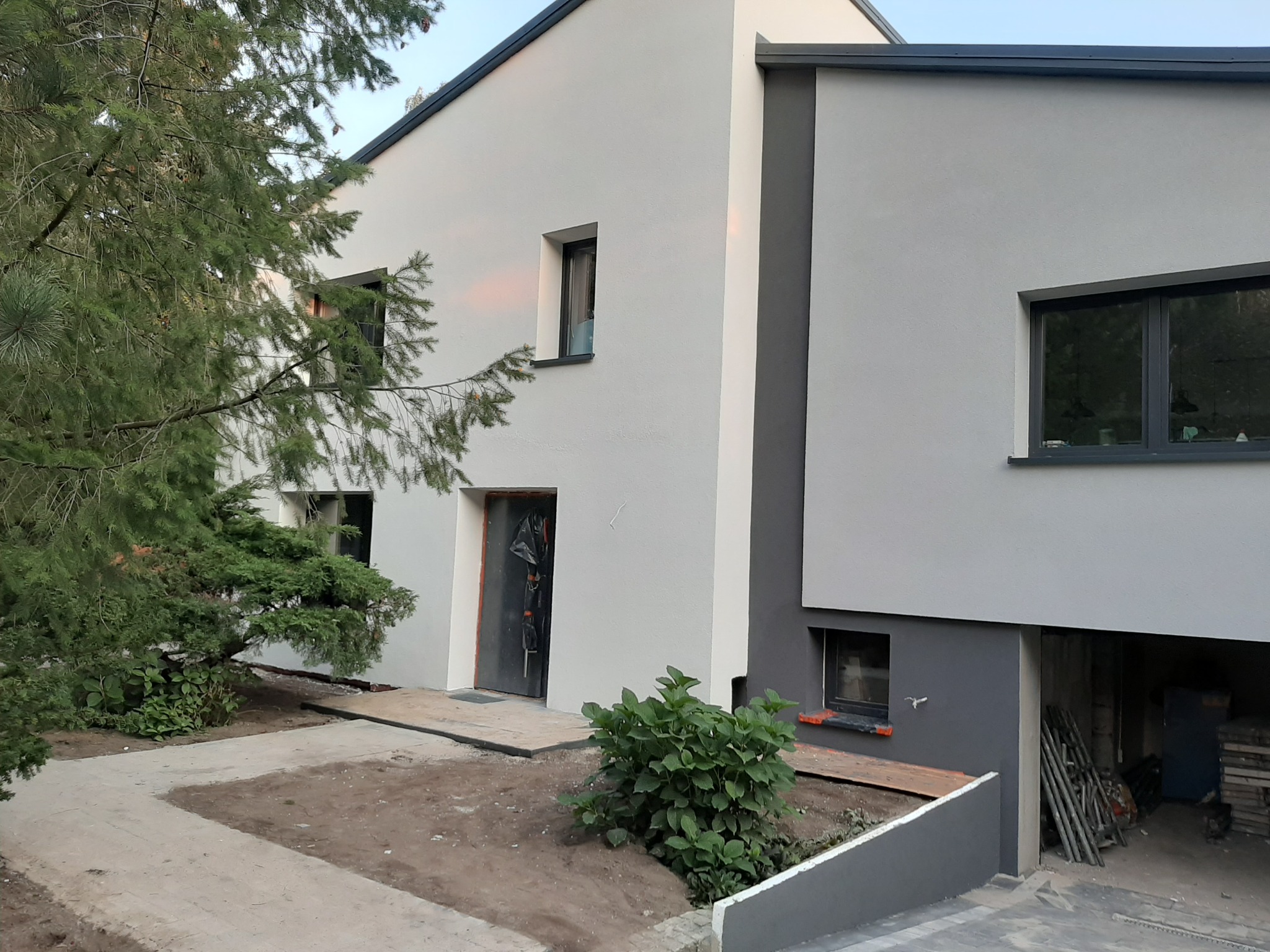 Nowoczesny, dwukondygnacyjny dom z jasnoszarą elewacją, ciemnoszarą stolarką okienną i drzwiami, widoczny fragment ogrodu z drzewem iglastym i utwardzonym podjazdem, stan surowy otwarty garażu.