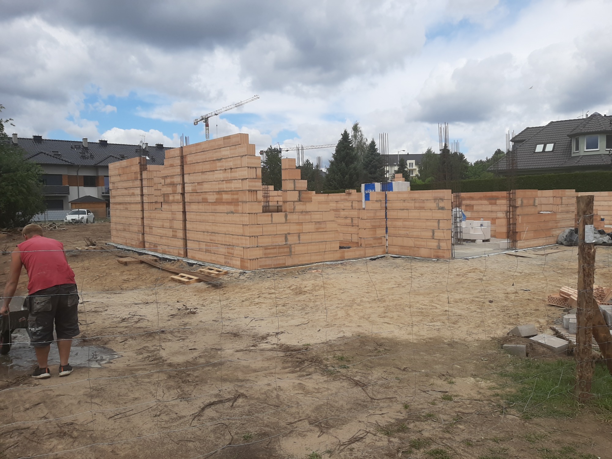 Wznoszenie ścian z bloczków na budowie domu jednorodzinnego, widoczny pracownik z taczką oraz zbrojenie pionowe w ścianach.