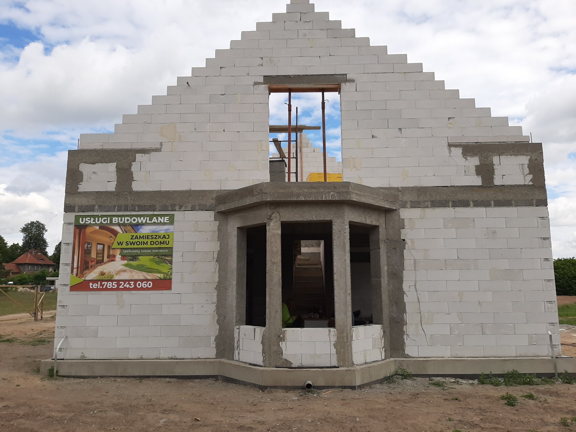 Surowy stan domu z białych bloczków, widoczne zbrojenia okienne i częściowo wymurowany wykusz, na ścianie reklama usług budowlanych.