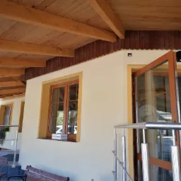 Elewacja budynku z drewnianym podbitkiem dachowym i oknami w drewnianych ramach, widoczne otwarte drzwi z naklejką na szybie i metalowa balustrada.