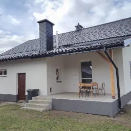 Nowoczesny dom jednorodzinny po remoncie elewacji, widoczny taras z drewnianymi elementami, grafitowa dachówka i komin, jasna elewacja z ciemną podmurówką, obok brązowe drzwi wejściowe.