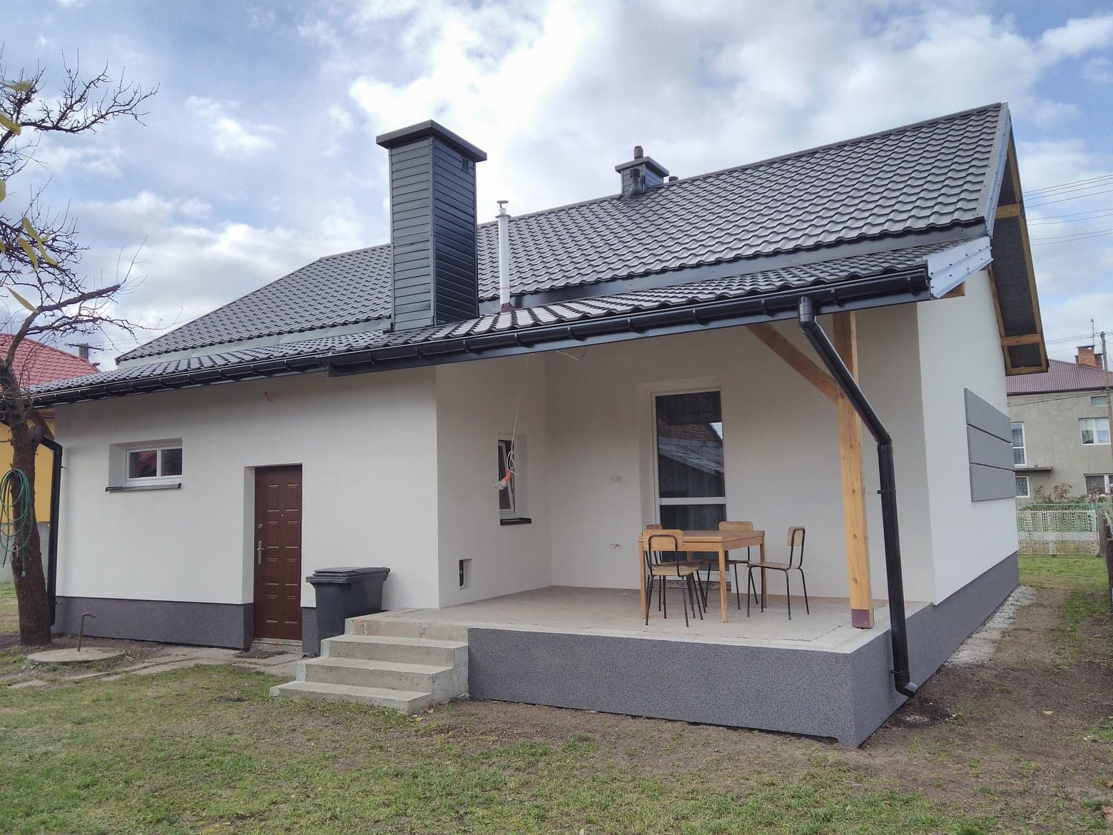 Nowoczesny dom jednorodzinny po remoncie elewacji, widoczny taras z drewnianymi elementami, grafitowa dachówka i komin, jasna elewacja z ciemną podmurówką, obok brązowe drzwi wejściowe.
