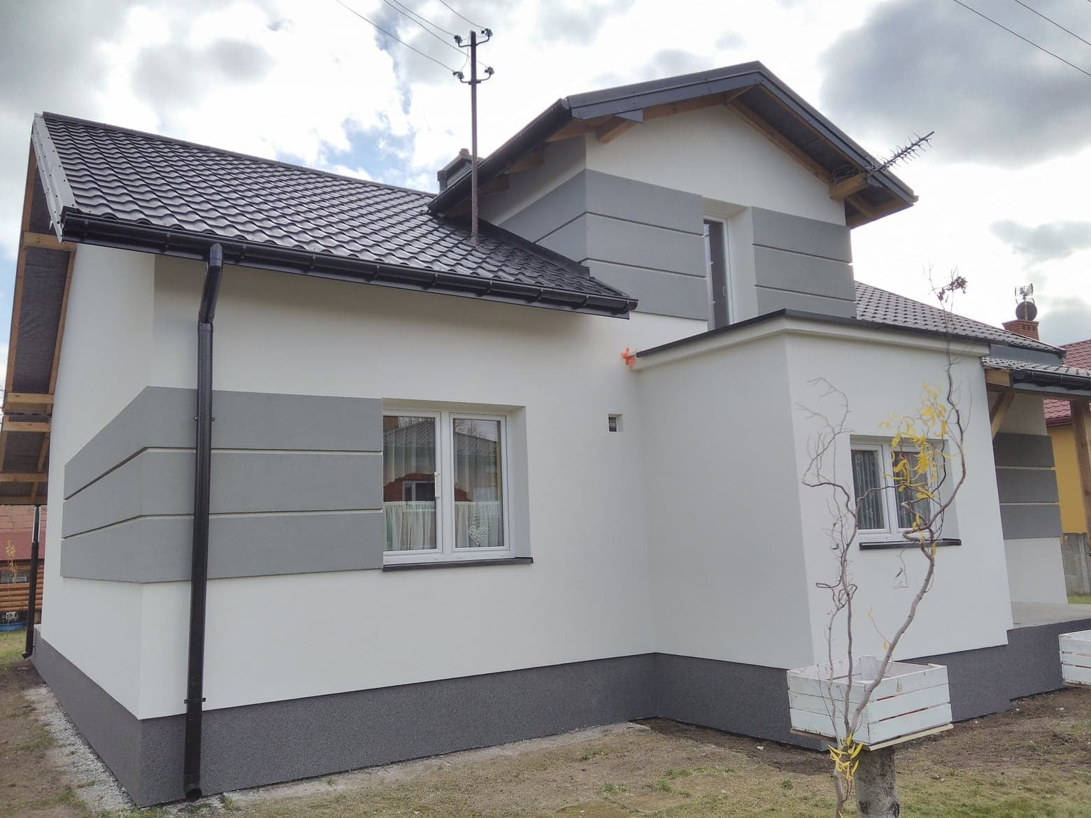 Elewacja domu jednorodzinnego z szarymi pasami dekoracyjnymi, czarną dachówką i rynnami, widok z zewnątrz.
