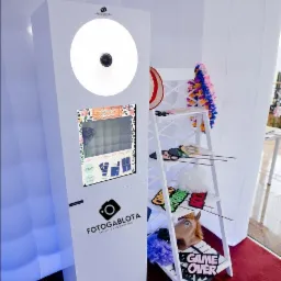 Biała fotobudka z ekranem dotykowym i oświetleniem LED, obok biały regał z rekwizytami do zdjęć: maski, peruki, hawajski naszyjnik, napis 'Game Over'.