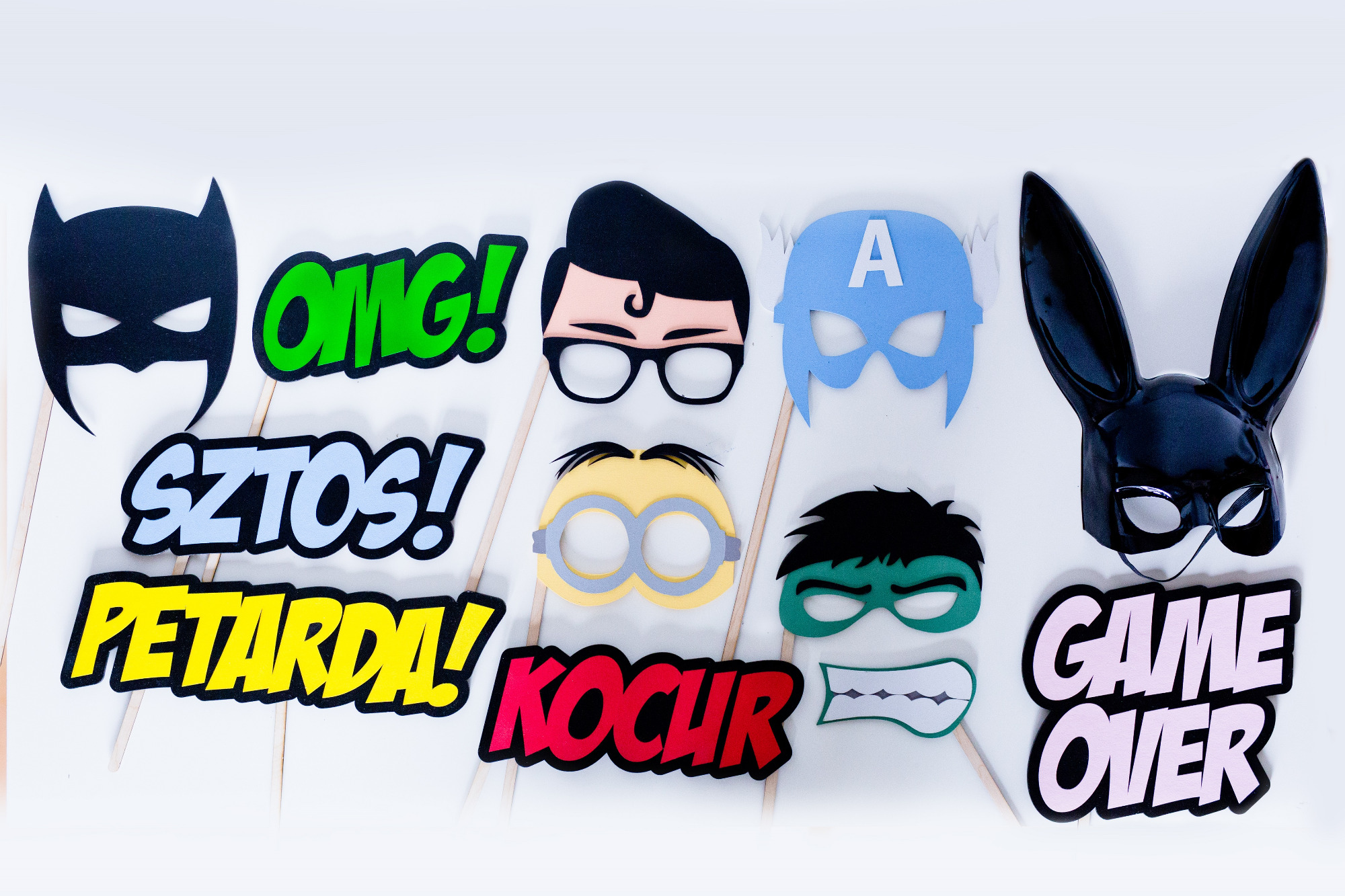 Różnorodne rekwizyty do fotobudki na patyku, w tym maski superbohaterów (Batman, Kapitan Ameryka), królicza maska, okulary Minionka oraz napisy 'OMG!', 'SZTOOS!', 'PETARDA!', 'KOCHUR' i 'GAME OVER'.