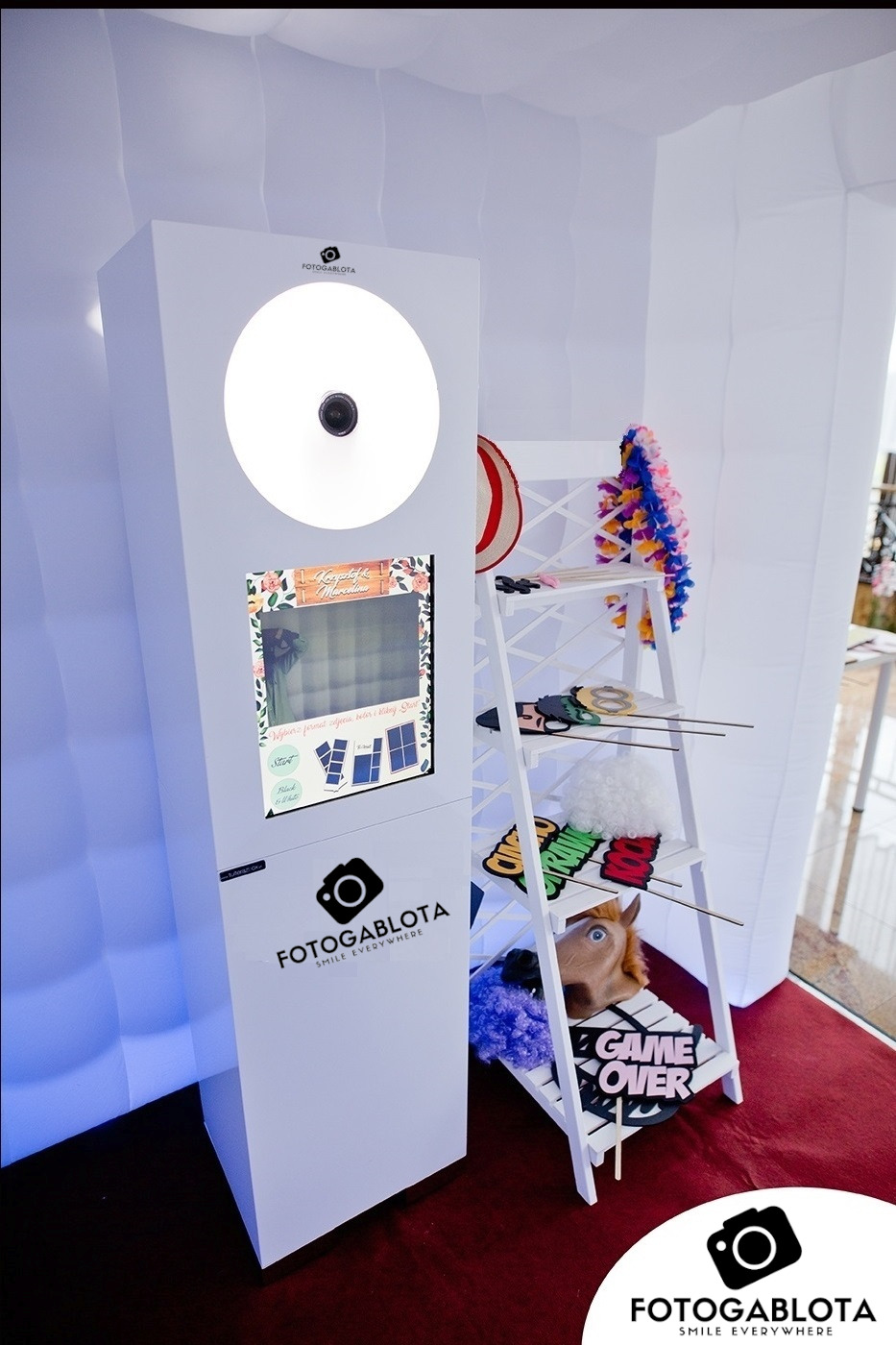 Biała fotobudka z ekranem dotykowym i oświetleniem LED, obok biały regał z rekwizytami do zdjęć: maski, peruki, hawajski naszyjnik, napis 'Game Over'.