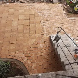 Widok z góry na częściowo ułożoną, pomarańczową kostkę brukową z zaokrąglonymi krawędziami, zagęszczarka obok bramy, narzędzia i ubrania na schodach.