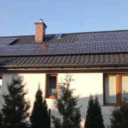 Panele fotowoltaiczne na dachu domu w Łazach, widoczne okna i komin z metalowym zakończeniem.