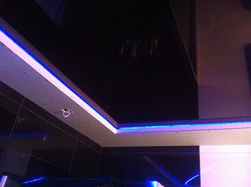 Błyszczący, czarny sufit podwieszany z oświetleniem LED w kolorze niebieskim, widoczne odbicia i fragmenty ścian.