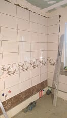 Ściana w trakcie kładzenia płytek ceramicznych w jasnych odcieniach, z dekoracyjnym pasem płytek z motywem roślinnym, widoczna drabina oparta o ścianę, surowy stan wykończenia pomieszczenia.