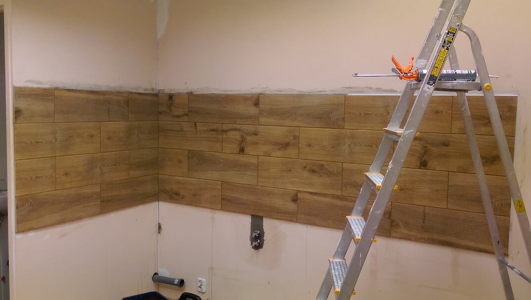 Układanie płytek imitujących drewno na ścianie, widoczna drabina i narzędzia do fugowania, etap prac wykończeniowych.
