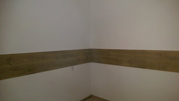 Fragment ściany wewnętrznej z dekoracyjną, drewnopodobną listwą przypodłogową na wysokości około metra, widoczna gładka, biała ściana i gniazdko elektryczne.