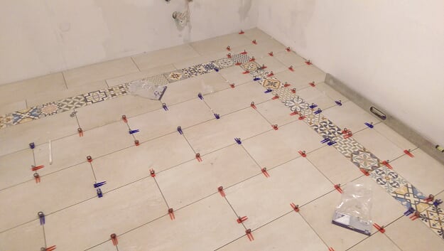 Układanie beżowych płytek podłogowych z dekoracyjnym pasem płytek wzorzystych, widoczne klipsy i kliny poziomujące, surowy stan ścian.