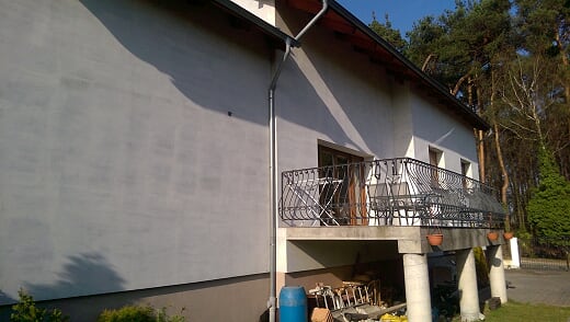 Dom jednorodzinny z nową, szarą elewacją i metalową rynną, widok na balkon z ozdobną balustradą i kolumnami, w tle las.