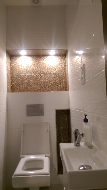 Nowoczesna łazienka z białymi płytkami, dekoracyjną wnęką z mozaiką i podświetleniem, wiszącą toaletą i białą umywalką z dozownikiem mydła.