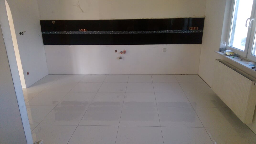 Wnętrze w trakcie remontu: białe płytki podłogowe, czarny panel dekoracyjny nad białą ścianą, widoczne instalacje elektryczne i hydrauliczne, grzejnik pod oknem.