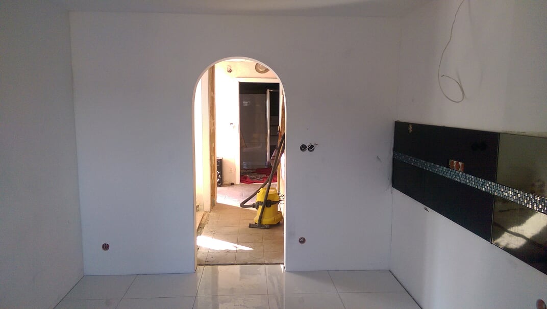 Wnętrze w trakcie remontu: widoczna surowa ściana z instalacją elektryczną, łukowate przejście do kolejnego pomieszczenia z odkurzaczem i fragmentem wykończonej ściany z płytkami