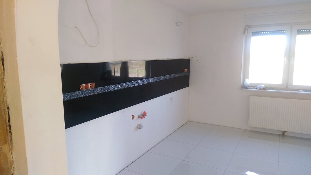 Częściowo wykończona kuchnia z czarnymi płytkami i mozaiką na ścianie, białe ściany i podłoga, widoczne instalacje wodne, okno z grzejnikiem.