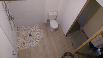 Wykończona łazienka z kabiną prysznicową wyłożoną dekoracyjnymi kamieniami i toaletą, widoczne prace wykończeniowe w sąsiednim pomieszczeniu.