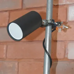 Czarna lampa punktowa z białym wnętrzem, zamocowana do metalowej rury na tle ceglanej ściany, widoczne złącza elektryczne.