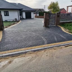 Nowo ułożony, ciemnoszary podjazd z kostki brukowej z żółtymi obrzeżami prowadzący do nowoczesnego domu z czarnym dachem i bramą wjazdową z szarym ogrodzeniem panelowym.