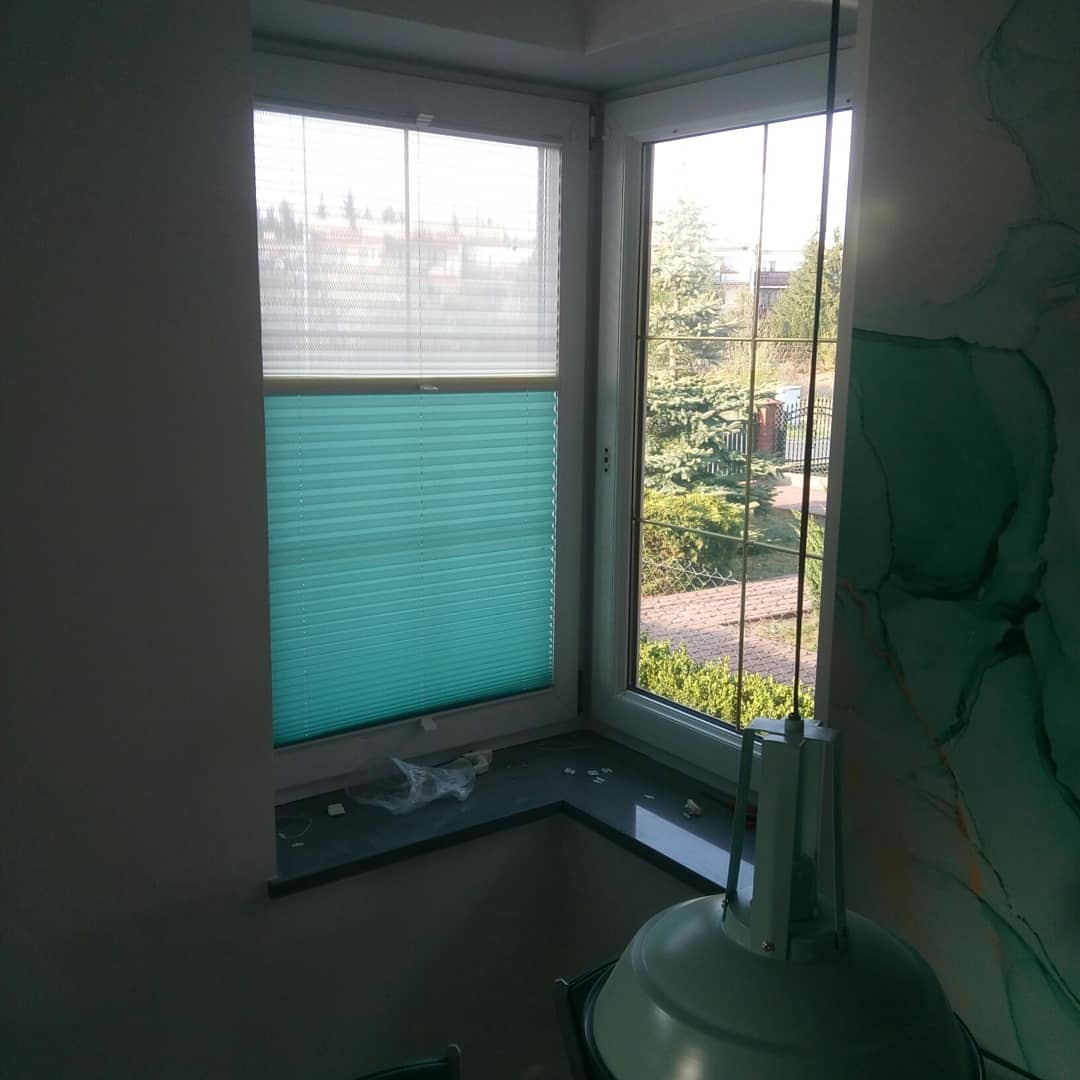 Okno PCV z zamontowaną turkusową roletą plisowaną od dołu i białą plisowaną od góry, widok na ogród.