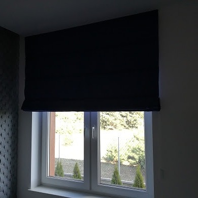 Zasłonięte okno z roletą rzymską w kolorze granatowym, widok na ogród z trzema miniaturowymi drzewkami iglastymi.