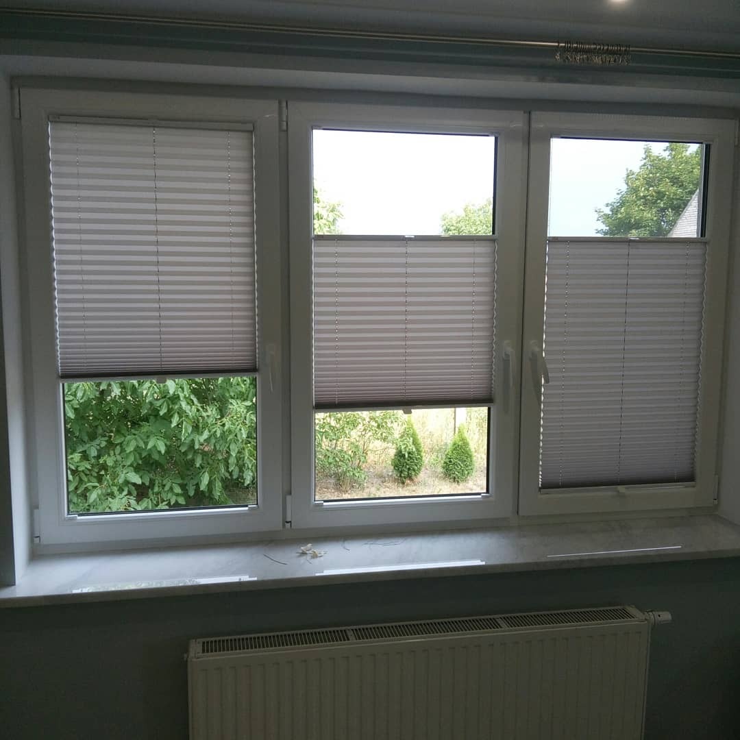 Trzyczęściowe okno z białymi ramami i zamontowanymi plisowanymi roletami w kolorze szarym, częściowo zasłoniętymi, widok na zieleń za oknem, parapet z marmurowym wzorem, grzejnik pod oknem.