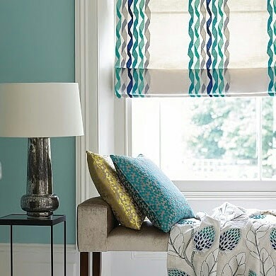 Pokój z turkusową ścianą, lampą na stoliku, poduszkami i roletą rzymską w niebiesko-szaro-zielone fale.