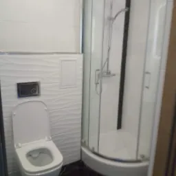 Nowoczesna łazienka z białą armaturą, kabiną prysznicową z zaokrąglonymi szybami i czarną marmurową podłogą, białe płytki na ścianach z subtelnym wzorem fal.