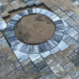 Układanie kostki brukowej wokół studzienki kanalizacyjnej, widoczne etapy prac i piasek zasypujący fugi.