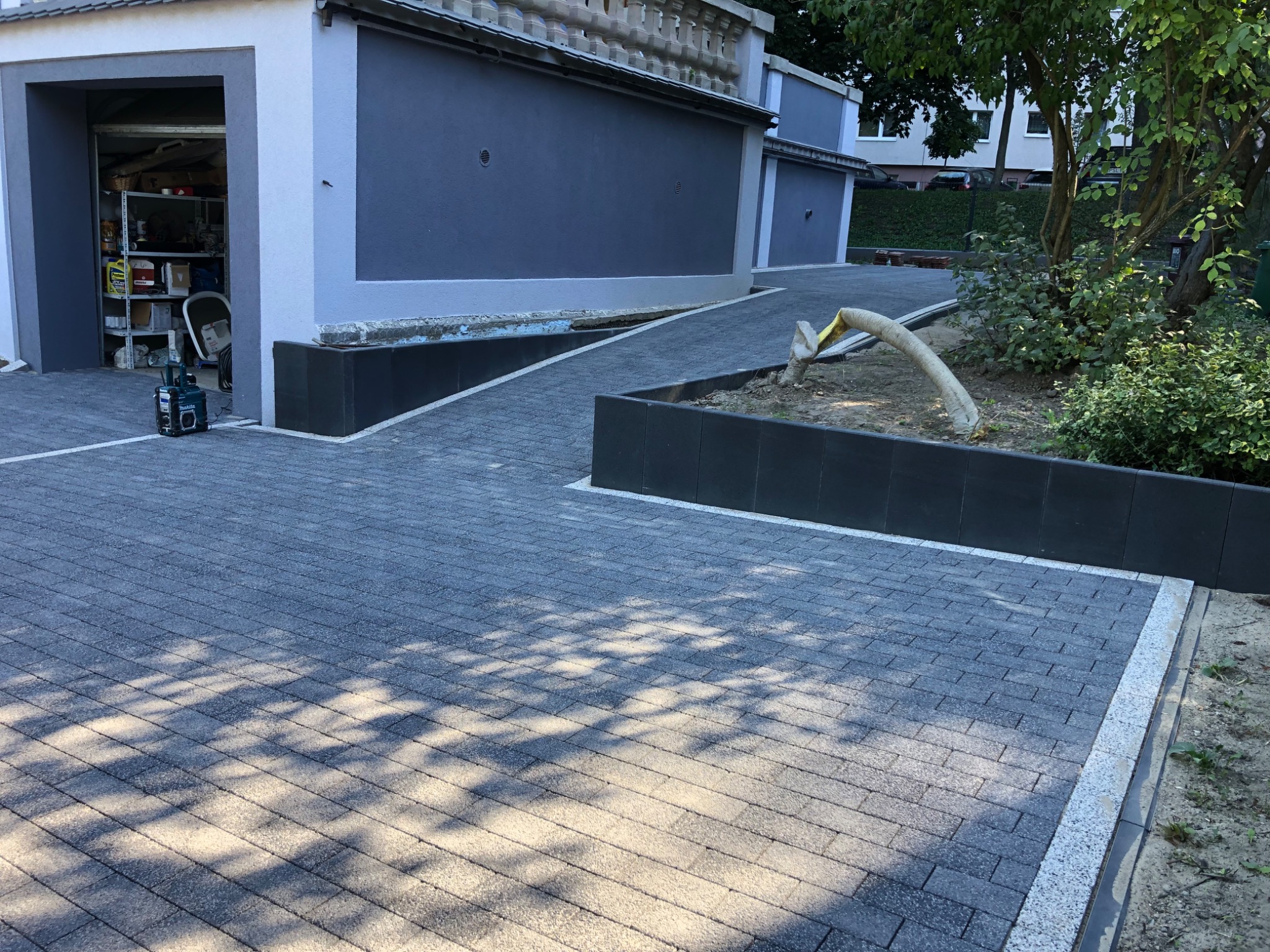 Nowo ułożona, szara kostka brukowa z białym obramowaniem, prowadząca do garażu i wzdłuż podwyższonej rabaty z czarnymi ściankami, częściowo zacieniona przez drzewa.