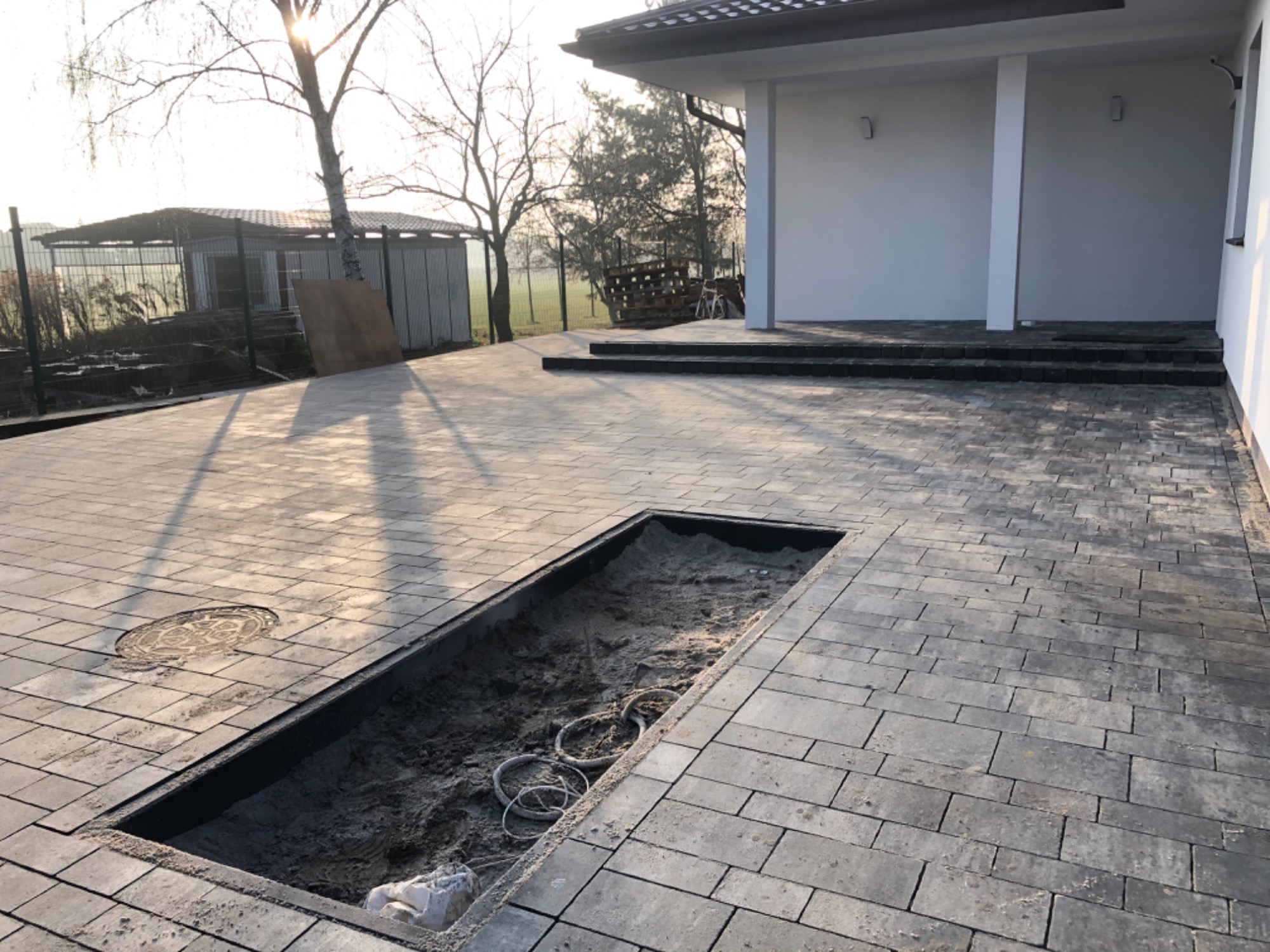 Nowoczesny podjazd z szarej kostki brukowej w trakcie budowy, z widocznym wykopem na instalacje, studzienką kanalizacyjną i budynkiem w tle.