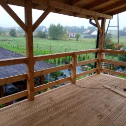 Zadaszony taras z drewnianą podłogą i balustradą, widok na ogród z altaną i zielony krajobraz w tle. Widać narzędzia pracy na podłodze tarasu.