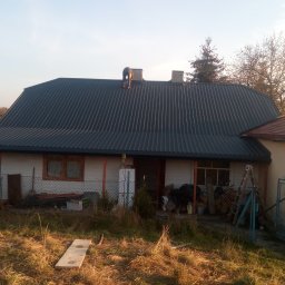 Tec-Dom - Pracownik na dachu drewnianego domu jednorodzinnego z blachy trapezowej, widoczne kominy i drzewo iglaste w tle.