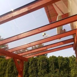 Tec-Dom - Zadaszenie tarasu z drewna i przezroczystych paneli, widok z dołu na konstrukcję i satelitę na dachu budynku w tle.