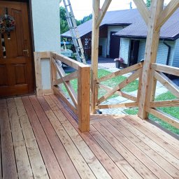 Tec-Dom - Nowo wybudowany taras z drewnianą podłogą i balustradą z krzyżowymi elementami, prowadzący do brązowych drzwi wejściowych domu, z widoczną drabiną opartą o ścianę w tle.